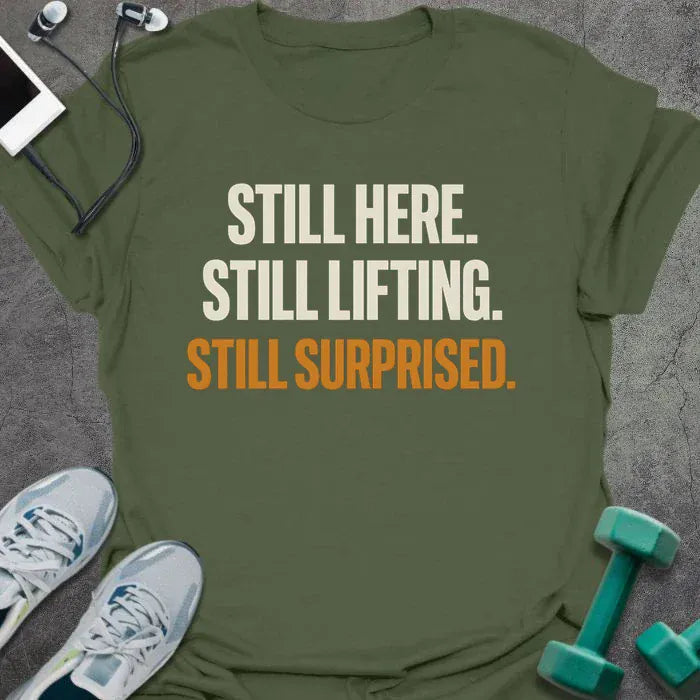 Military green t-shirt printed with "Still Here. Still Lifting. Still Surprised." in white and orange