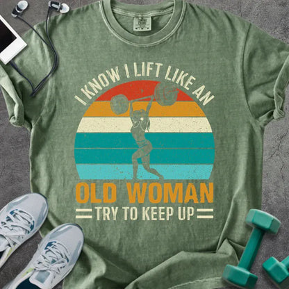 Moss t-shirt printed with "I Know I Lift Like an Old Woman Try to Keep Up" in orange and multicolor, with a lifting silhouette and rainbow sunset