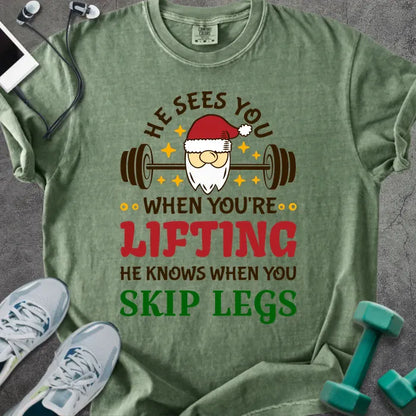 Moss t-shirt printed with "He Sees You When You're Lifting He Knows When You Skip Legs" in multicolor, with Santa holding a barbell
