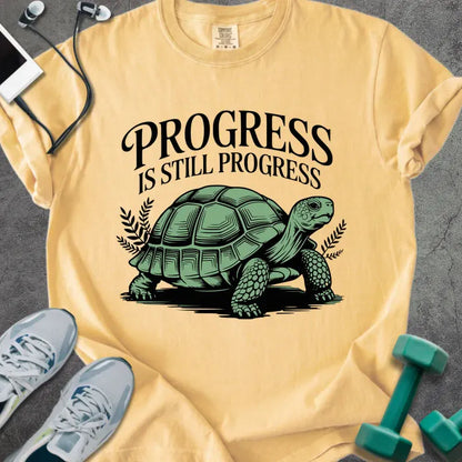 Mustard t-shirt printed with "Progress is Still Progress" in green, with a turtle illustration and decorative leaves