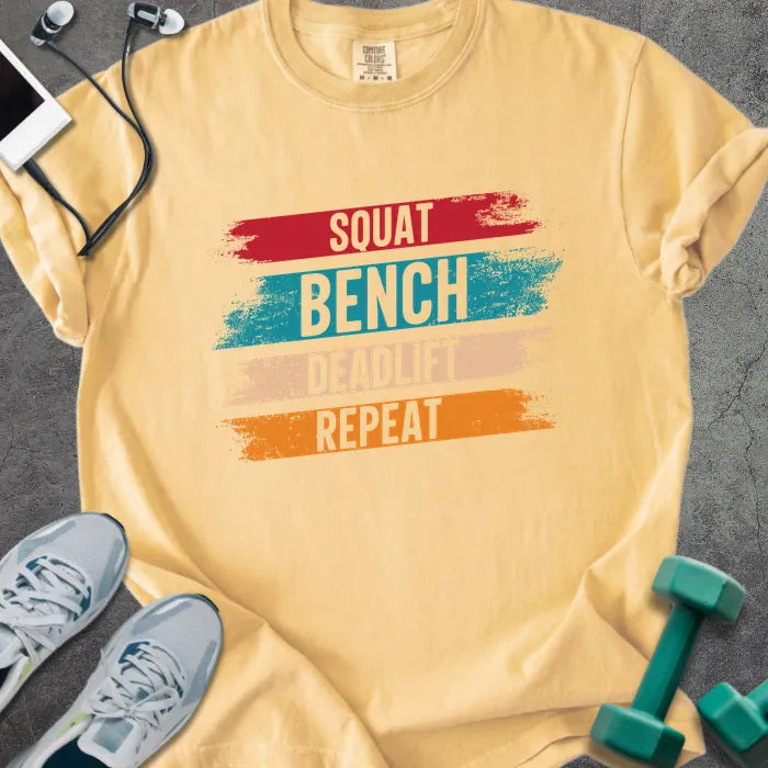 Mustard t-shirt printed with "Squat Bench Deadlift Repeat" in red, teal, tan, orange, with red, teal, tan, and orange paint-stroke bands behind the words