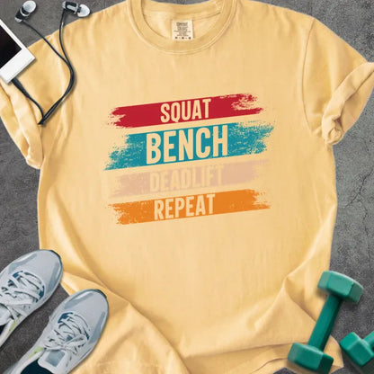 Mustard t-shirt printed with "Squat Bench Deadlift Repeat" in red, teal, tan, orange, with red, teal, tan, and orange paint-stroke bands behind the words