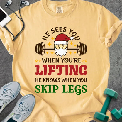 Mustard t-shirt printed with "He Sees You When You're Lifting He Knows When You Skip Legs" in multicolor, with Santa holding a barbell