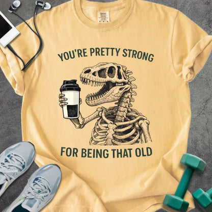 Mustard t-shirt printed with "You're Pretty Strong for Being That Old" in green, with skeleton holding a coffee cup
