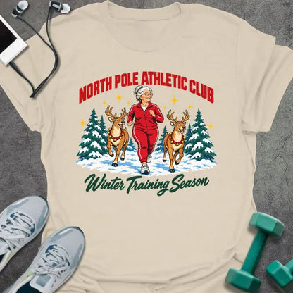 Natural t-shirt printed with "NORTH POLE ATHLETIC CLUB" in red and "Winter Training Season" in green script, with a white-haired woman in a red tracksuit jogging between two reindeer through snowy pine trees