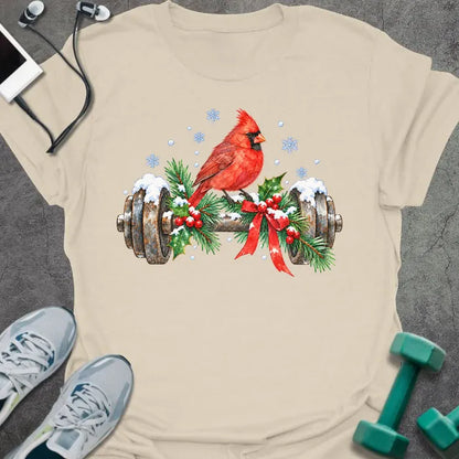 Natural t-shirt printed with a watercolor red cardinal perched on a snowy dumbbell trimmed with holly, pine branches and a red bow, with blue snowflakes