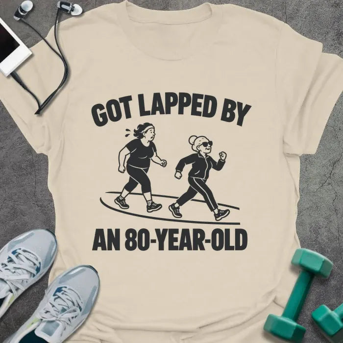 Natural t-shirt printed with "GOT LAPPED BY AN 80-YEAR-OLD" in black, with a tired jogger being passed by an older woman in sunglasses speed walking on a track