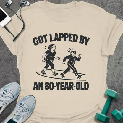 Natural t-shirt printed with "GOT LAPPED BY AN 80-YEAR-OLD" in black, with a tired jogger being passed by an older woman in sunglasses speed walking on a track