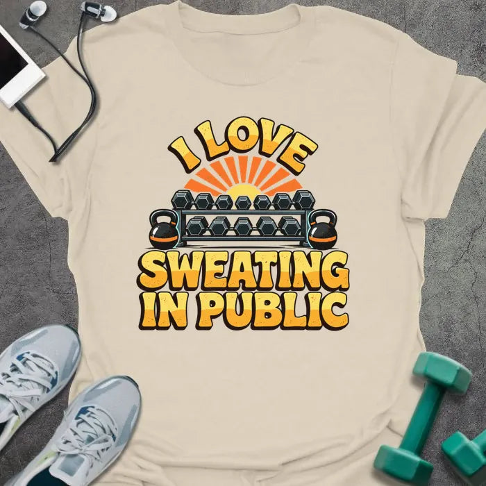 Natural t-shirt printed with "I LOVE SWEATING IN PUBLIC" in yellow and orange, with a dumbbell rack, kettlebells and a retro sunset