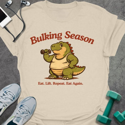 Cream t-shirt printed with "Bulking Season Eat. Lift. Repeat. Eat Again." in brown and green, with dinosaur lifting weights graphic