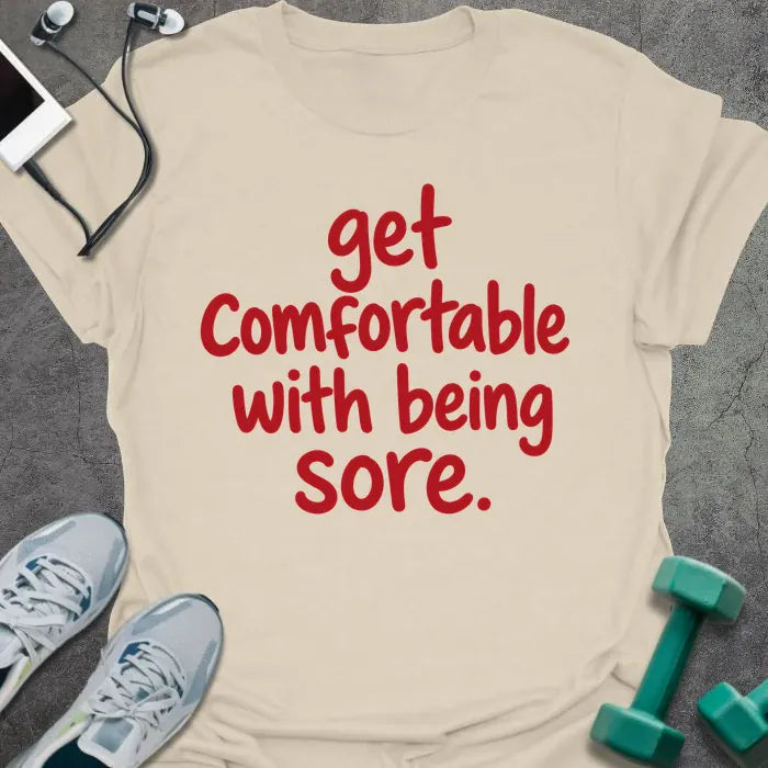 Natural t-shirt printed with "get comfortable with being sore." in red