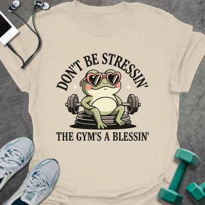 Cream t-shirt printed with "Don't Be Stressin' the Gym's a Blessin'" in red, with an illustration of a frog with heart-shaped sunglasses lifting dumbbells