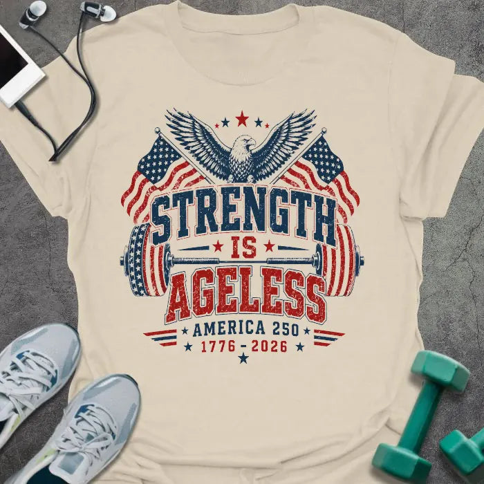 Cream t-shirt printed with "Strength is Ageless America 250 1776 - 2026" in red and blue, with an eagle, American flags, and dumbbell