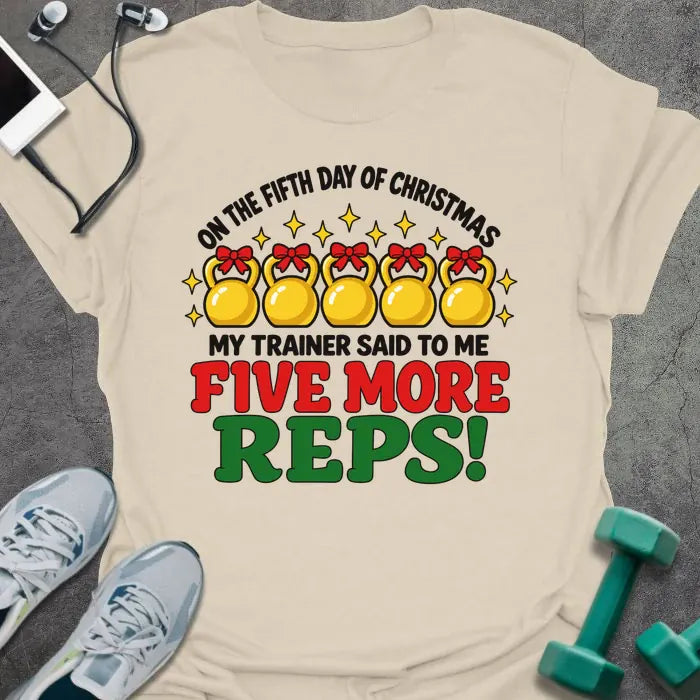 Natural t-shirt printed with "ON THE FIFTH DAY OF CHRISTMAS MY TRAINER SAID TO ME, FIVE MORE REPS!" in black, red and green, with five gold kettlebells tied with red bows