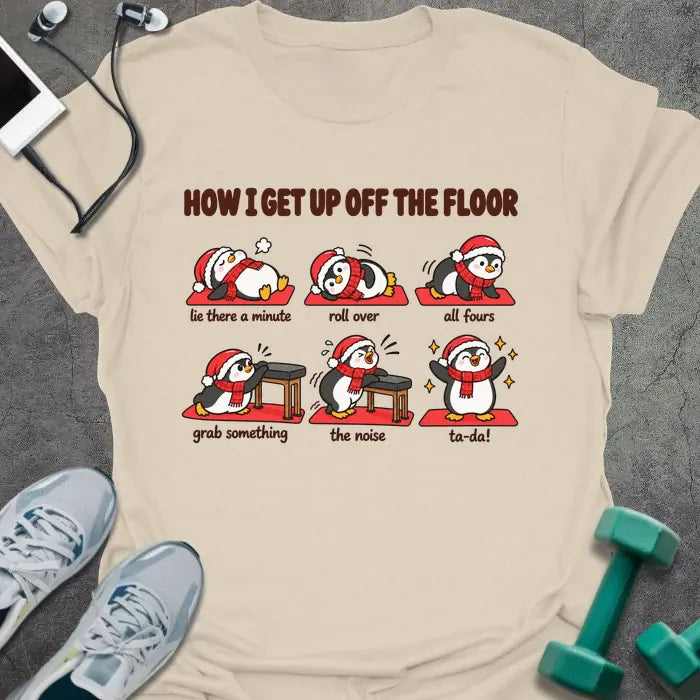 Natural t-shirt printed with "HOW I GET UP OFF THE FLOOR" in red, with six penguins in Santa hats showing the steps: lie there a minute, roll over, all fours, grab something, the noise, ta-da!