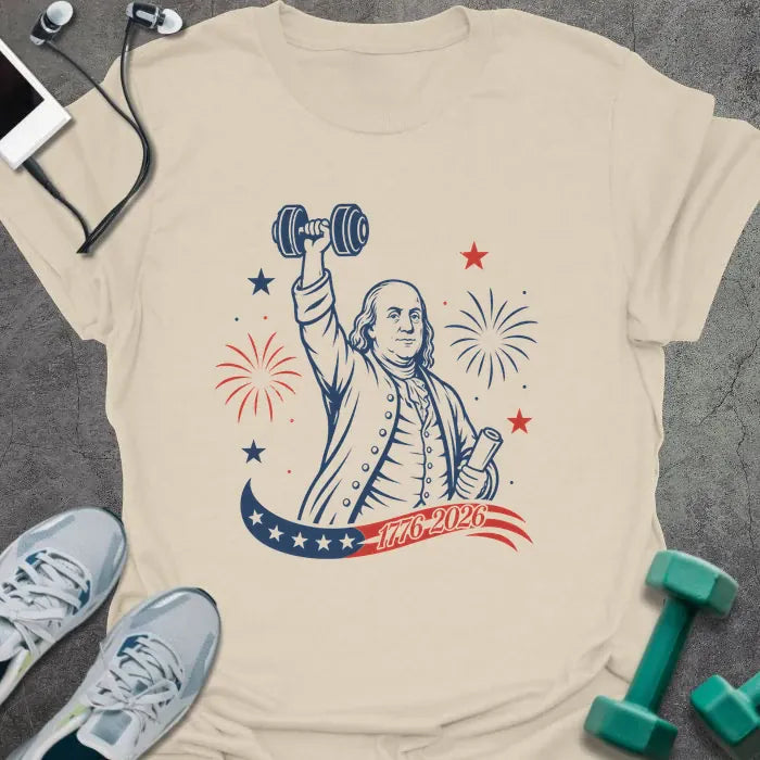 Cream t-shirt printed with "1776-2026" in navy, red, with Benjamin Franklin holding a dumbbell overhead and a scroll, fireworks and stars