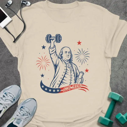 Cream t-shirt printed with "1776-2026" in navy, red, with Benjamin Franklin holding a dumbbell overhead and a scroll, fireworks and stars