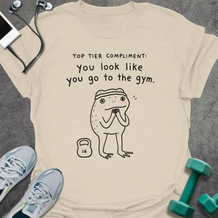 Natural t-shirt printed with "TOP TIER COMPLIMENT: you look like you go to the gym." in black, with frog graphic holding barbells