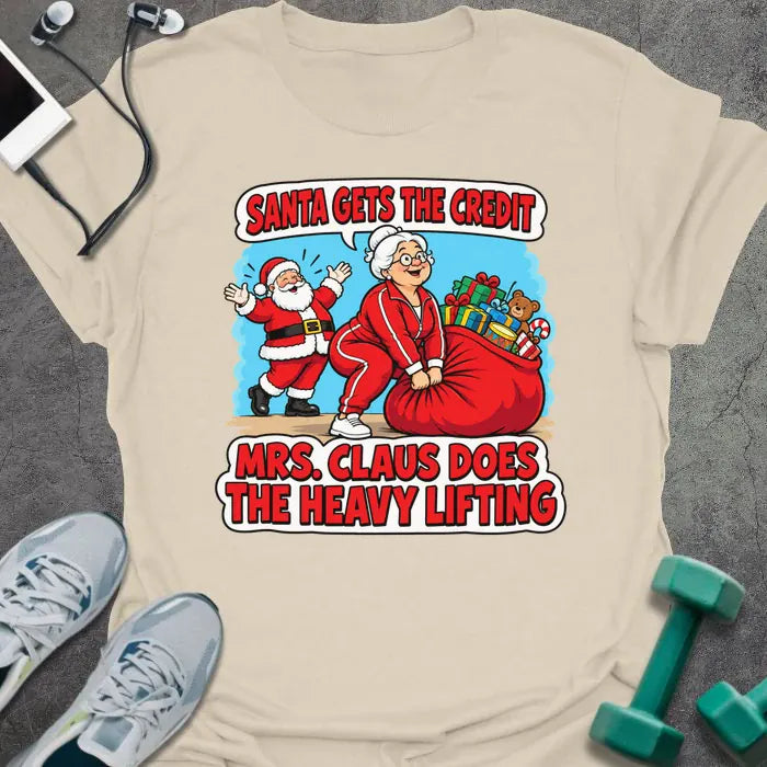 Natural t-shirt printed with "SANTA GETS THE CREDIT, MRS. CLAUS DOES THE HEAVY LIFTING" in red and white, with Mrs. Claus hauling a sack of gifts while Santa waves