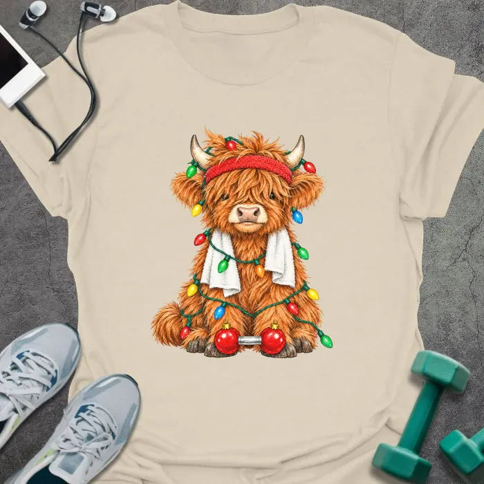 Natural t-shirt printed with a fluffy highland cow in a red headband and towel, tangled in Christmas lights beside a barbell with ornament plates