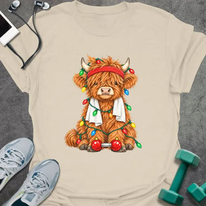 Natural t-shirt printed with a fluffy highland cow in a red headband and towel, tangled in Christmas lights beside a barbell with ornament plates