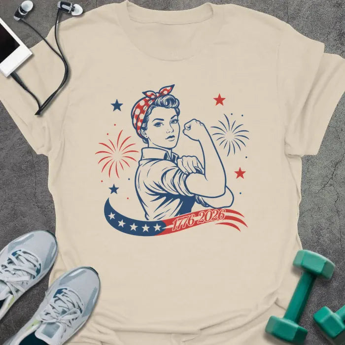 Cream t-shirt printed with "1776-2026" in navy, red, with Rosie the Riveter flexing her bicep among fireworks and stars