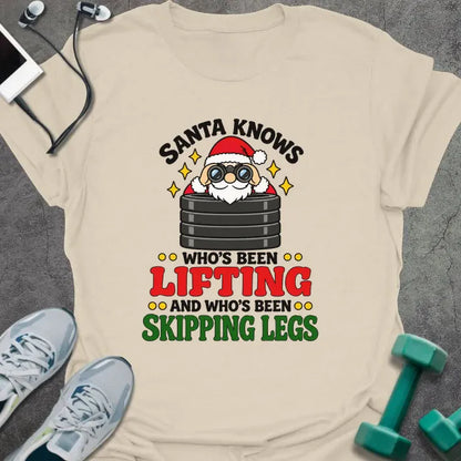Natural t-shirt printed with "SANTA KNOWS WHO'S BEEN LIFTING AND WHO'S BEEN SKIPPING LEGS" in black, red and green, with Santa peering through binoculars over a stack of weight plates