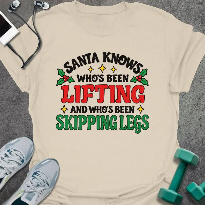 Natural t-shirt printed with "SANTA KNOWS WHO'S BEEN LIFTING AND WHO'S BEEN SKIPPING LEGS" in black, red and green, with holly and gold stars