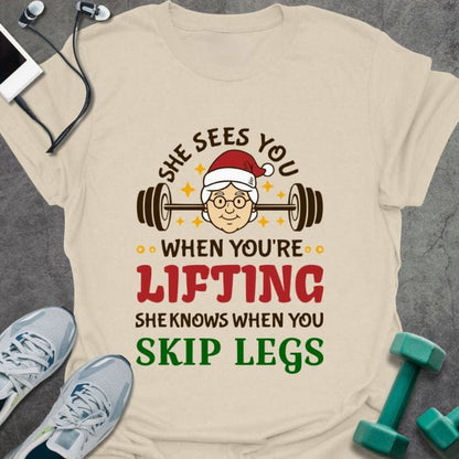 Cream t-shirt printed with "She Sees You When You're Lifting Sheknows When You Skip Legs" in black, red, and gold, with female Santa and barbell