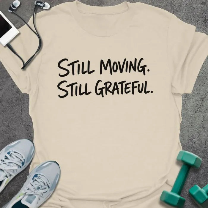 Natural t-shirt printed with "Still Moving. Still Grateful." in black handwritten lettering