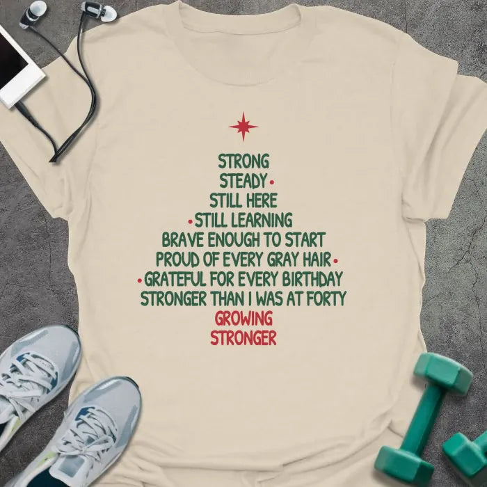 Natural t-shirt printed with a Christmas tree made of words, "STRONG, STEADY, STILL HERE, STILL LEARNING, BRAVE ENOUGH TO START, PROUD OF EVERY GRAY HAIR, GRATEFUL FOR EVERY BIRTHDAY, STRONGER THAN I WAS AT FORTY, GROWING STRONGER", in green and red with a red star on top