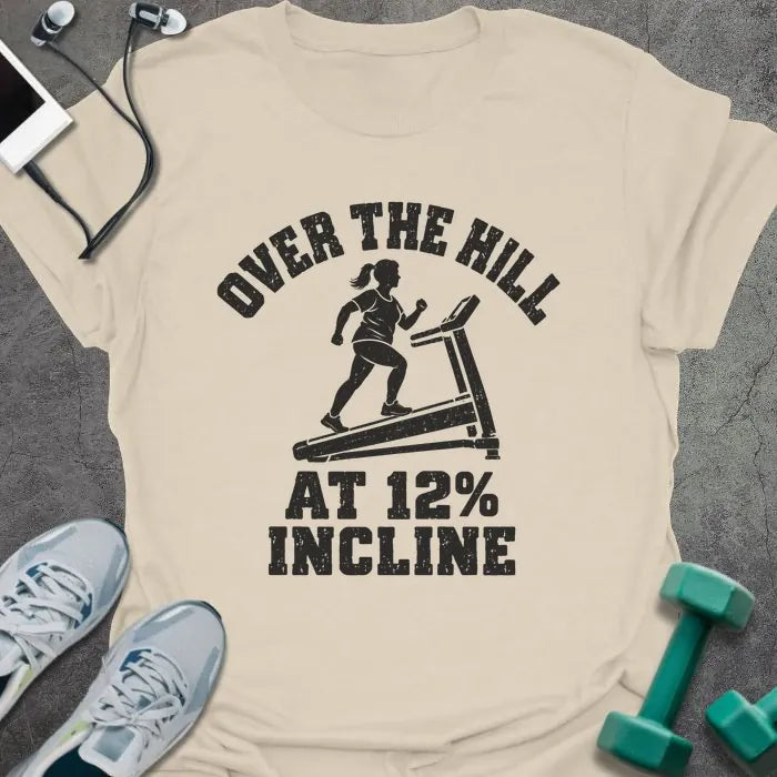 Natural t-shirt printed with "OVER THE HILL AT 12% INCLINE" in charcoal, with a woman walking on a treadmill