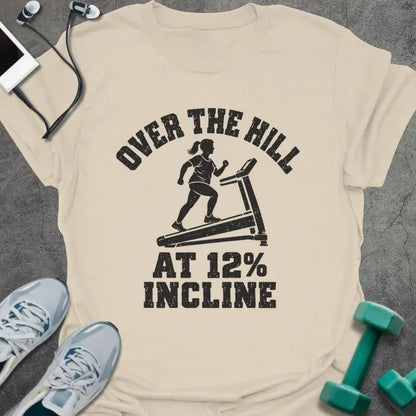 Natural t-shirt printed with "OVER THE HILL AT 12% INCLINE" in charcoal, with a woman walking on a treadmill