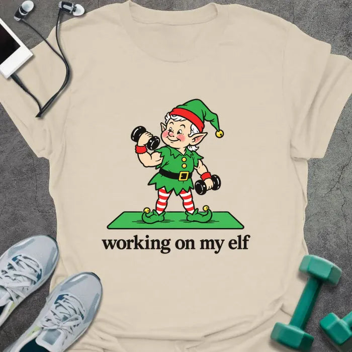 Natural t-shirt printed with "working on my elf" in black, with a vintage elf lifting dumbbells on a green mat