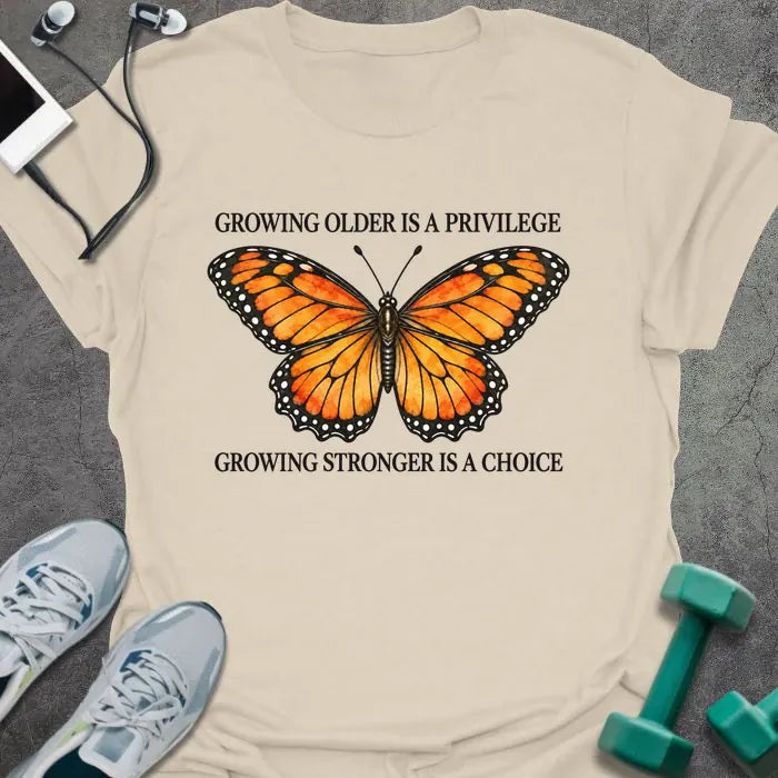 Natural t-shirt printed with "GROWING OLDER IS A PRIVILEGE, GROWING STRONGER IS A CHOICE" in black, with an orange watercolor monarch butterfly