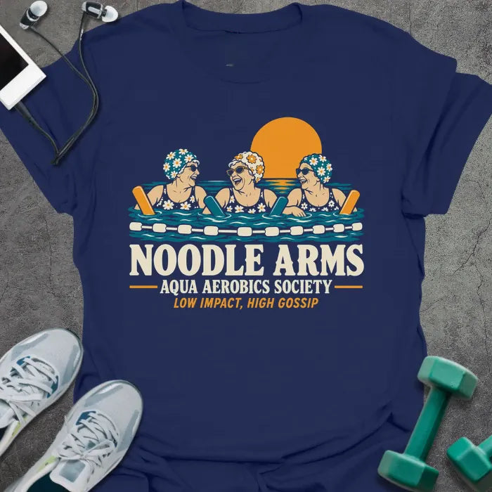 Navy t-shirt printed with "NOODLE ARMS, AQUA AEROBICS SOCIETY, LOW IMPACT, HIGH GOSSIP" in cream and orange, with three older women in flowered swim caps and sunglasses laughing in a pool with pool noodles at sunset