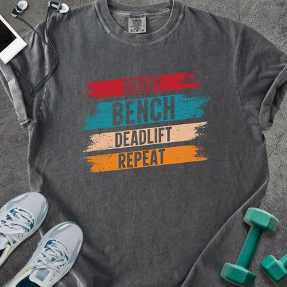 Pepper t-shirt printed with "Squat Bench Deadlift Repeat" in red, teal, tan, orange, with red, teal, tan, and orange paint-stroke bands behind the words
