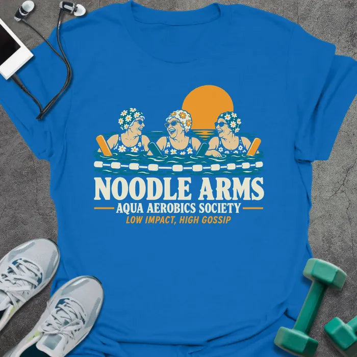 Royal blue t-shirt printed with "NOODLE ARMS, AQUA AEROBICS SOCIETY, LOW IMPACT, HIGH GOSSIP" in cream and orange, with three older women in flowered swim caps and sunglasses laughing in a pool with pool noodles at sunset