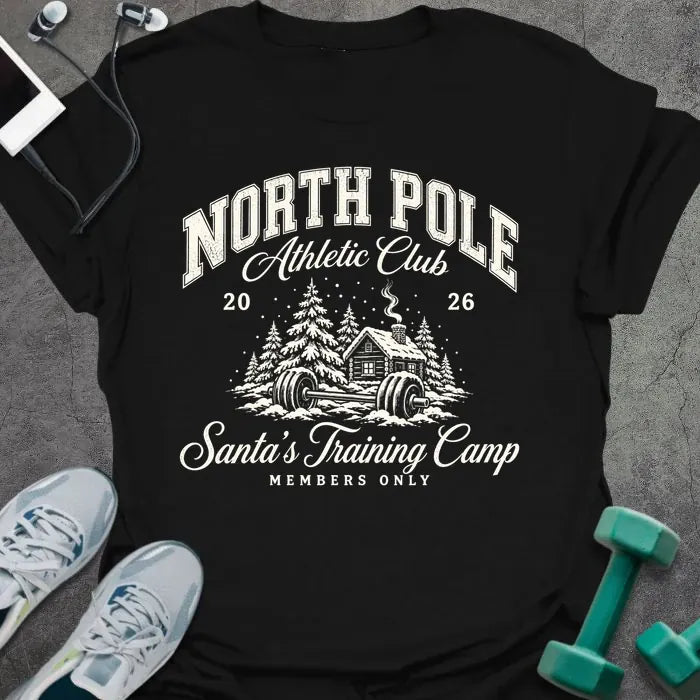 Black t-shirt printed with "NORTH POLE Athletic Club 2026, Santa's Training Camp, MEMBERS ONLY" in cream, with snowy pine trees, a log cabin and a barbell in the snow