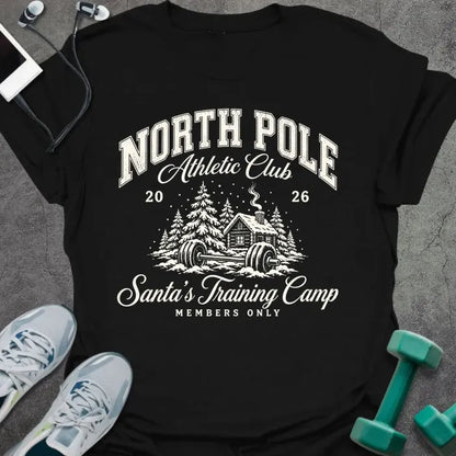 Black t-shirt printed with "NORTH POLE Athletic Club 2026, Santa's Training Camp, MEMBERS ONLY" in cream, with snowy pine trees, a log cabin and a barbell in the snow