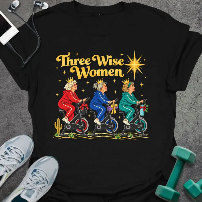 Black t-shirt printed with "Three Wise Women" in gold, with three older women in crowns and red, blue and teal tracksuits riding spin bikes under a bright star