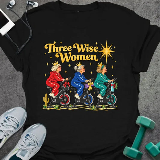 Black t-shirt printed with "Three Wise Women" in gold, with three older women in crowns and red, blue and teal tracksuits riding spin bikes under a bright star