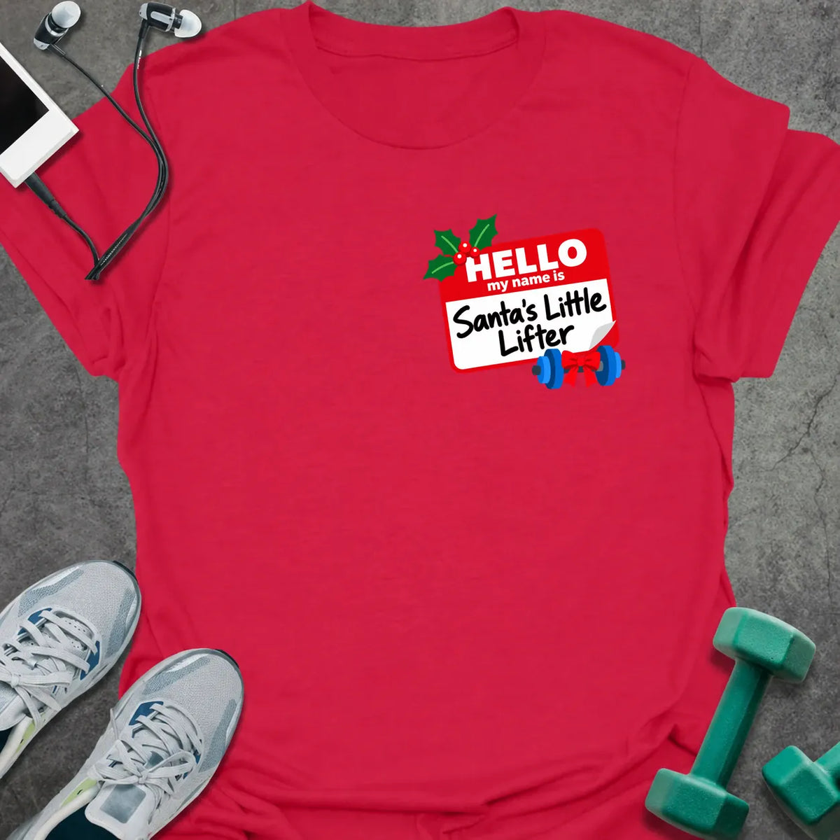 Cherry red t-shirt printed with a small red name tag on the chest reading "HELLO my name is Santa's Little Lifter", with holly and a blue dumbbell tied with a red bow