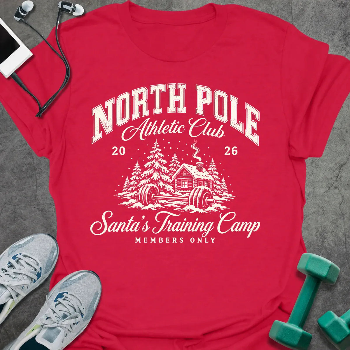 Cherry red t-shirt printed with "NORTH POLE Athletic Club 2026, Santa's Training Camp, MEMBERS ONLY" in cream, with snowy pine trees, a log cabin and a barbell in the snow