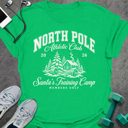 Irish green t-shirt printed with "NORTH POLE Athletic Club 2026, Santa's Training Camp, MEMBERS ONLY" in cream, with snowy pine trees, a log cabin and a barbell in the snow