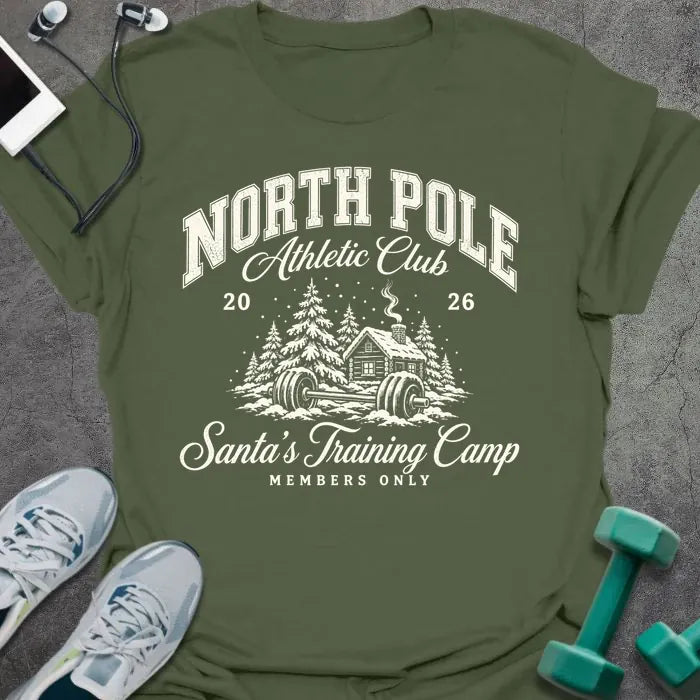 Military green t-shirt printed with "NORTH POLE Athletic Club 2026, Santa's Training Camp, MEMBERS ONLY" in cream, with snowy pine trees, a log cabin and a barbell in the snow