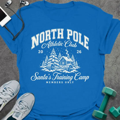 Royal blue t-shirt printed with "NORTH POLE Athletic Club 2026, Santa's Training Camp, MEMBERS ONLY" in cream, with snowy pine trees, a log cabin and a barbell in the snow