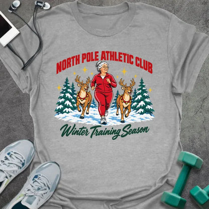 Sport grey t-shirt printed with "NORTH POLE ATHLETIC CLUB" in red and "Winter Training Season" in green script, with a white-haired woman in a red tracksuit jogging between two reindeer through snowy pine trees