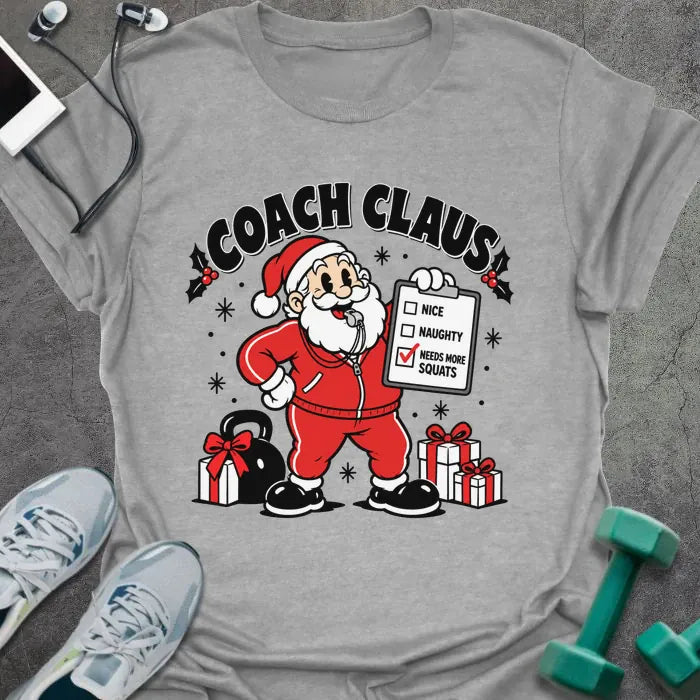 Sport grey t-shirt printed with "COACH CLAUS" in black, with a cartoon Santa in a red tracksuit and whistle holding a clipboard with "NEEDS MORE SQUATS" checked, next to a kettlebell and wrapped presents