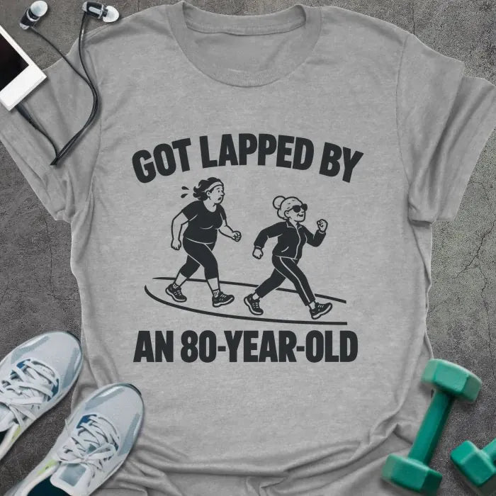 Sport grey t-shirt printed with "GOT LAPPED BY AN 80-YEAR-OLD" in black, with a tired jogger being passed by an older woman in sunglasses speed walking on a track